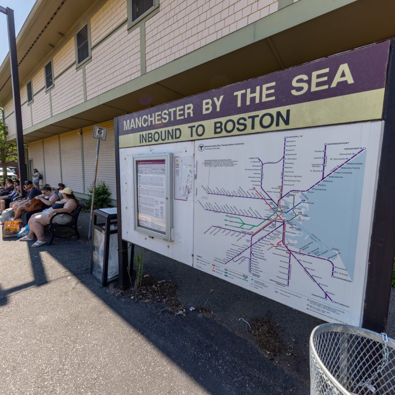Manchester Station inbound platform on the Newburyport/Rockport Commuter Rail Line. People waiting for the train after a day at the beach. Sign shows station name and Commuter Rail system map.
